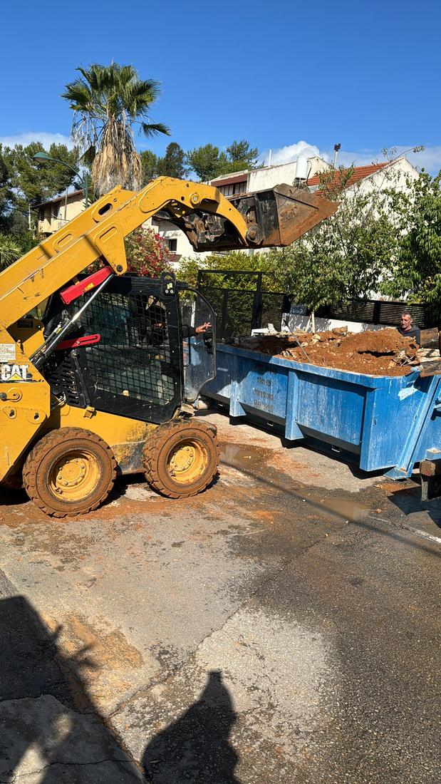בובקט CAT צהוב מעמיס אדמה לתוך מכולה כחולה ברחוב
