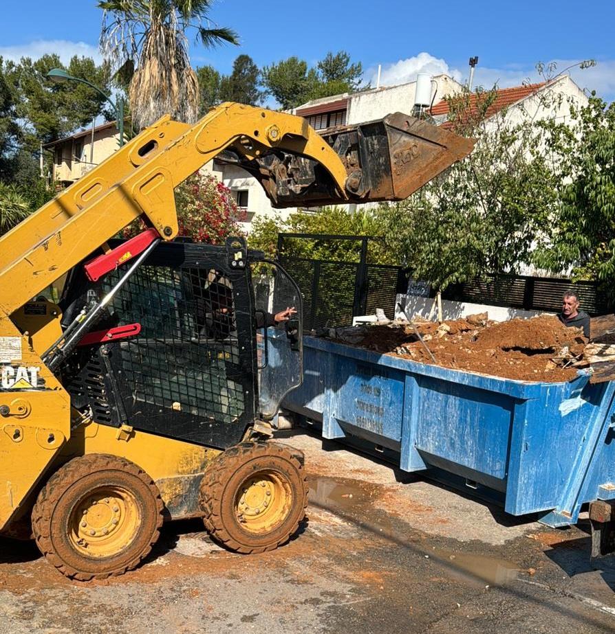 בובקט CAT מרים כף אדמה מעל מכולה כחולה בחצר פרטית