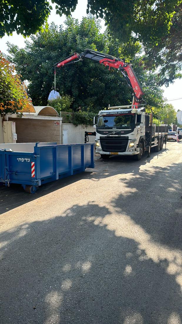 משאית מנוף וולוו מעבירה מטען מעל מכולה כחולה ברחוב שקט
