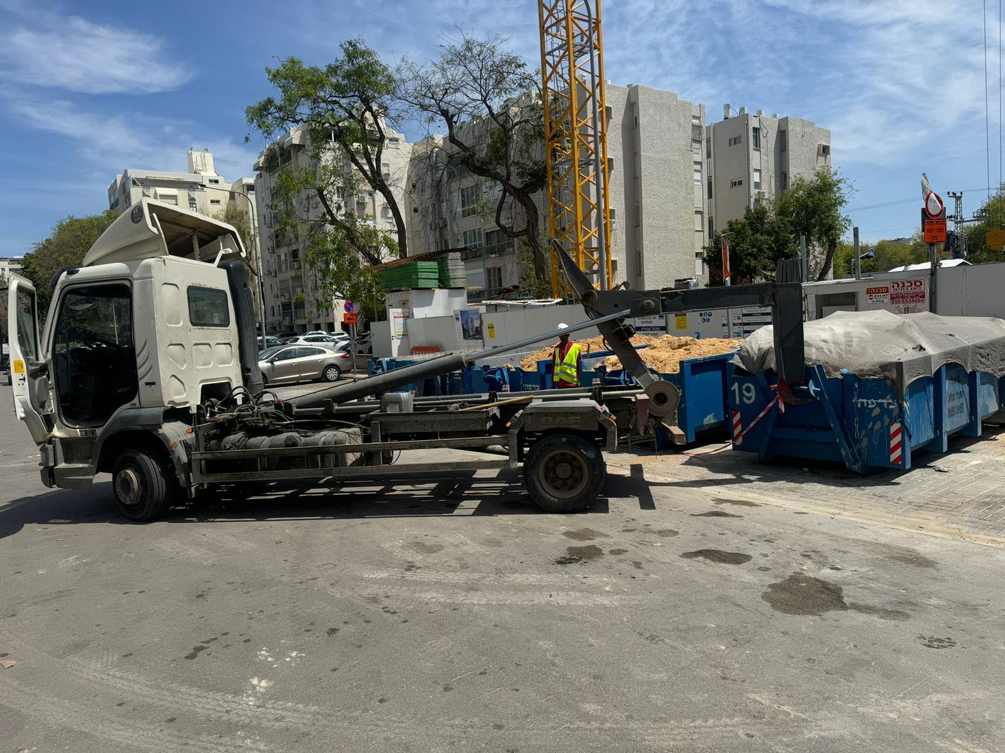 משאית רול־און לבנה מציבה מכולה באתר בנייה עם עגורן ברקע