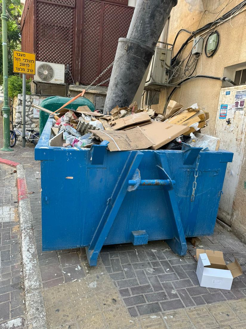 מכולה כחולה מלאה בקרטונים ואריזות על מדרכה עירונית